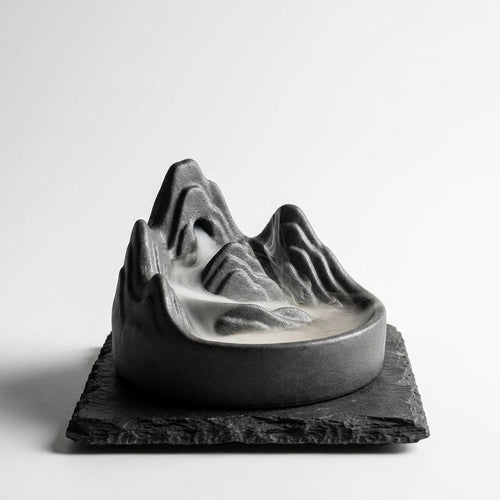 Dark stoneware backflow incense burner with white smoke cascading like mountain fog
