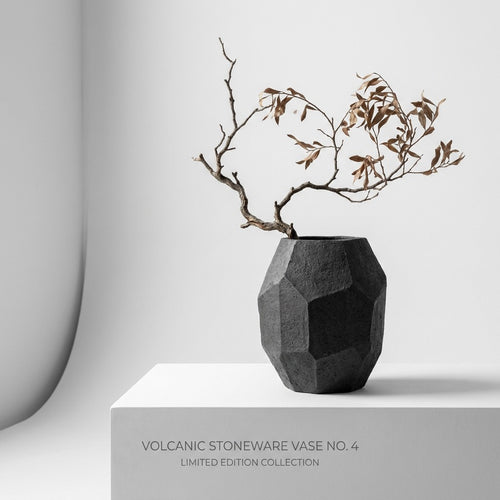 Textured brutalist dark stone vase holding single dried branch on concrete surface
